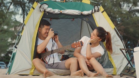 young loving couple pretty while camp taking photo by camera while sitting at the camping tent on beach in holiday vacation time at sunset, young asian group woman and man in summer travel outdoor