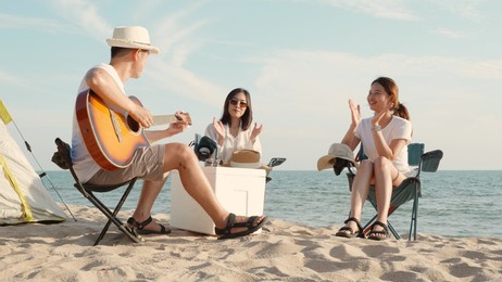 happy friend have fun playing guitar and clap in camp they smiling together in holiday on sand beach near camping tent vacation time at sunset, young asian group woman and man in summer travel outdoor