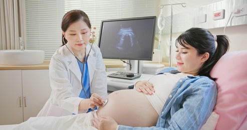 asian female doctor exams a pregnant woman lying in hospital ward by stethoscope