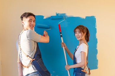 young asian couple decorate home - they painting wall with blue color together and smiling at you