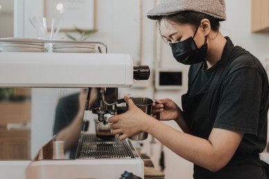 asian woman barista using coffee machine in coffee shop. female barista wearing medical face mask working in coffee cafe.