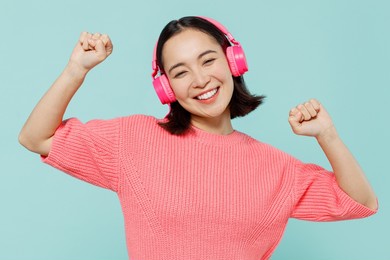 young smiling happy woman of asian ethnicity 20s wearing pink sweater headphones listen to music dance isolated on pastel plain light blue color background studio portrait. people lifestyle concept