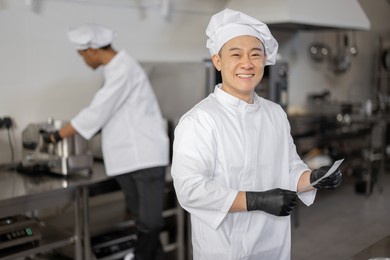 portrait of smiling asian chef in uniform standing with printed order in professional kitchen with latin guy cooking on background