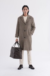 fashion model. young man with hairstyle in striped coat with khaki  pants holding handbag posing in studio on white background


