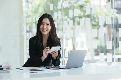 portrait of asian business woman paying bills online with laptop in office. beautiful girl with computer and chequebook, happy paying bills. startup business financial calculate account concept