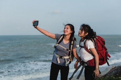 two asian backpacker women are taking selfies with their smartphones at the seaside.