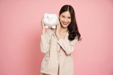 portrait of happy asian businesswoman holding white piggy bank isolated on pink background, saving money and financial economize concept