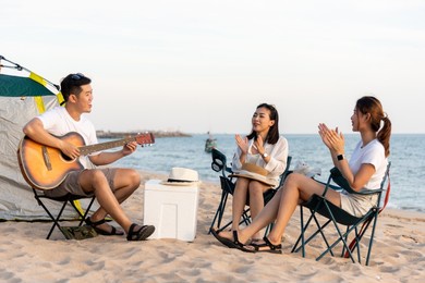 happy friend have fun playing guitar and clap in camp they smiling together in holiday on sand beach near camping tent vacation time at sunset, young asian group woman and man in summer travel outdoor