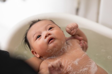 close up an adorable baby boy with  wet body in bath tub, his mother's hands pouring water on him. drops of water and bubbles soap flying around is breast. the kid likes shower time with mom.