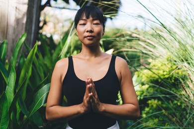 cropped image of asian girl practicing yoga in upward salute pose outdoor. harmony and mental health. young thoughtful woman with closed eyes wearing sportswear in garden on bali island. sunny day