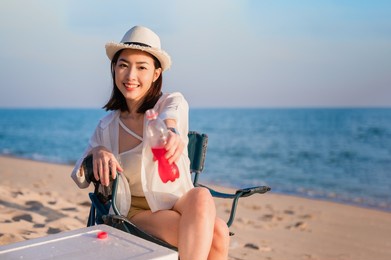 happy beautiful asian woman sitting on picnic chair wear sunglasses and hat holding red beverage bottle on beach in outdoor vacation summer. young female smile relax and drink cocktail travel camping.