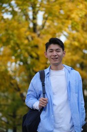 asian male college student and blurred yellow ginkgo biloba tree in autumn. carrying a shoulder bag and smiling at camera