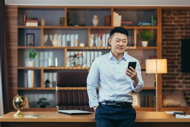 successful asian businessman in modern home office, looks at phone screen, smiles and rejoices, portrait of successful freelancer in shirt, uses phone app