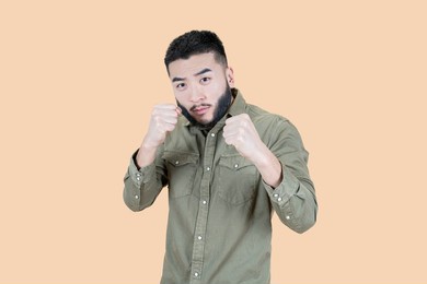 boxing and self defense. portrait of bearded asian man in casual clothes holding clenched fists, ready to defend himself in fight. isolated studio shot on beige background.