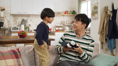 happy asian father hugging his cute son while he covers his eyes and gives him a surprise present in living room at home. father’s day celebration concept