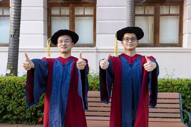 two happy proud phd graduated male students in academic dress gown show a thumb up
