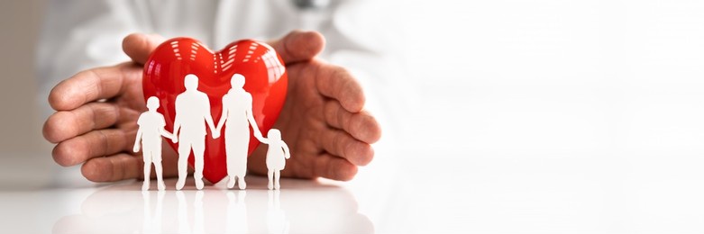 doctor's hand protecting red heart with family figure on reflective desk