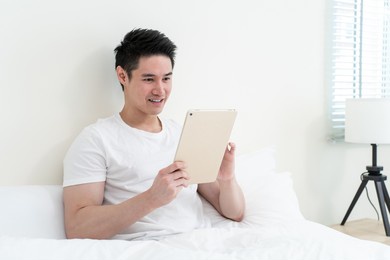 asian handsome man read online news from tablet in morning at home. happy attractive male lying down on bed in bedroom enjoy holiday weekend, using smartphone communication with technology in house.