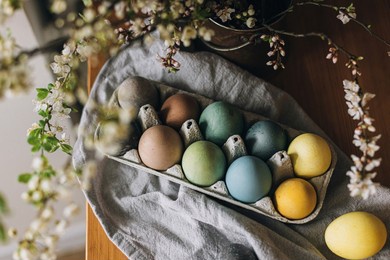 happy easter! easter eggs on rustic table with cherry blossoms. natural dyed colorful eggs in paper tray on wooden board with linen napkin and spring flowers. countryside still life