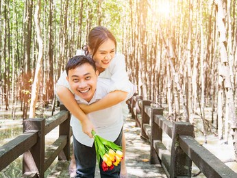 an asian woman in a white dress holding a bouquet of flowers happily rides on the back of an asian man in the mangrove forest. to show love and happiness after being proposed with cool shade