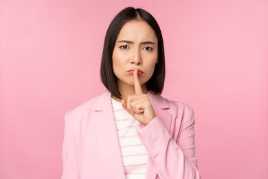hush, taboo concept. portrait of asian businesswoman showing shush gesture, shhh sign, press finger to lips, standing over pink background in suit