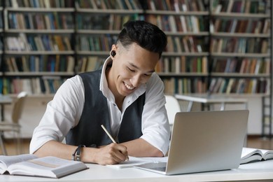 handsome asian guy hold pencil take notes in copybook while studying alone in library. student wear earbuds listen audio task, makes assignment sit at table. education, improve knowledge, tech concept