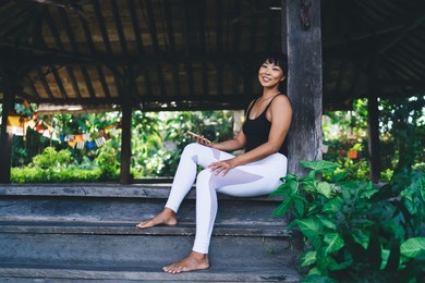 asian girl with smartphone resting on stairs on wooden terrace. tourism, vacation and weekend. young smiling beautiful brunette woman looking at camera. idyllic and tranquil lifestyle on bali island