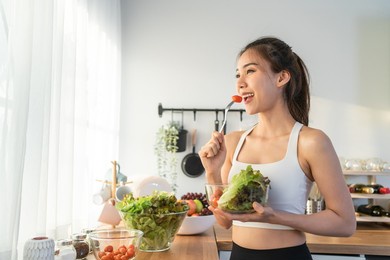 asian attractive sport woman holding salad bowl and eat vegetables. beautiful sport girl in sportswear enjoy eat clean vegetables after exercise for health in house. diet and healthy food concept.