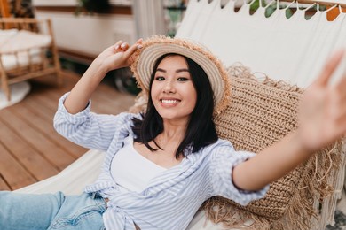 smiling young asian woman lying in hammock near camper van, taking selfie on camping trip in countryside. cool young lady making mobile photo of herself on summer vacation outdoors