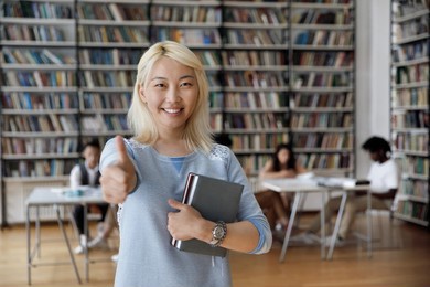asian student girl with blond dyed hair hold books pose in library showing thumb up at cam, mates on background. high school, university education, knowledge and studentship, excellent studies concept