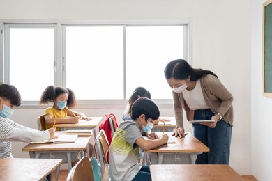 asian female teacher teaching her school kids. concentrated diverse ethnicity small children wearing protective face mask sitting at the desk in classroom, writing.