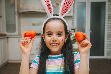 a little girl with pink bunny ears holds easter eggs on her face near her eyes in the kitchen.
