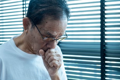 senior asian man coughing wearing glasses, after waking up in the morning, in the bedroom, wearing a white shirt, covering his mouth with hands