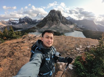 handsome asian male traveler taking selfie portrait on the hill with mount assiniboine in national park at bc, canada