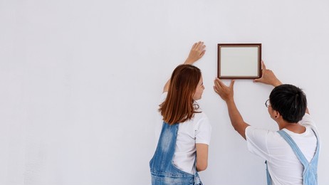 portrait of asian couple in bear suit  put picture frame on white wall in new apartment, renovation concept 