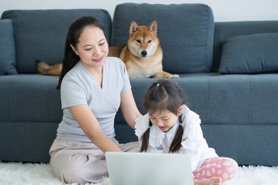 mother and daughter in the living room with a shiba inu sitting on the sofa. asian mom and little girl look at laptop and shiba dog stay on sofa
