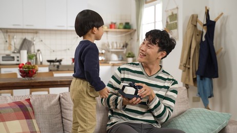 adorable asian toddler boy covering his dad’s eyes and showing him a gift from behind on father’s day. the father opening the box with smile on sofa