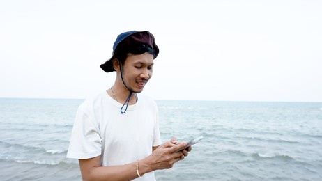 good-looking asian man, standing by the sea on the phone playing in the evening with clear air, cool breeze, happy smiling face.