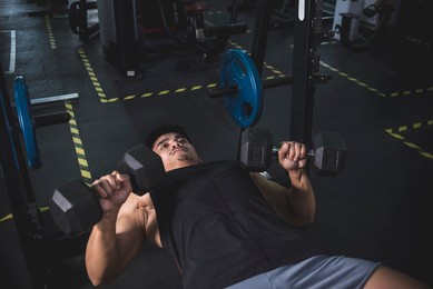 a muscled and fit asian man does a set of dumbbell bench presses. working out and training chest muscles at the gym. wearing a dark tank top.