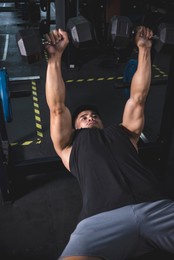 a muscled and fit asian man does a set of dumbbell bench presses. working out and training chest muscles at the gym. wearing a dark tank top.