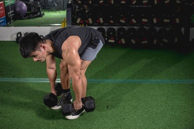 a buff and muscled asian man picks up a pair of dumbbells for a set of bent-over rows. weight and resistance training at the gym.