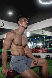 a shirtless asian man with a well built chest and a tight small waist sitting on a bench. a jacked male in his late 20s with an aesthetic physique at the gym. wearing gray shorts.