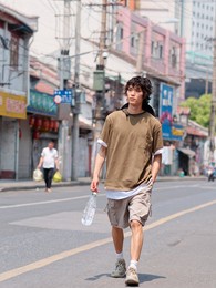 portrait of handsome chinese young man in brown t-shirt and pants walking on shanghai old town street with bottle of water in hand, front view of cool young man.