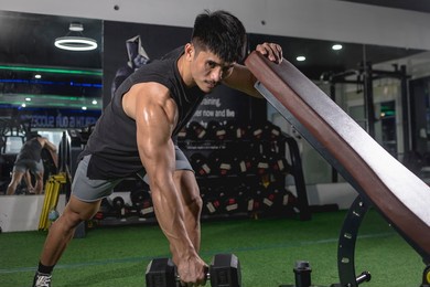 a handsome and fit asian guy does one-arm dumbbell rows while leaning on an incline bench. performing negative reps. back exercise variation. weight and resistance training at the gym.