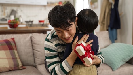 cute asian preschool boy surprising father with a gift on father’s day. smiling asian dad feeling touched and hugging his baby son in the living room at home
