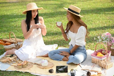 two gorgeous asian women having lunch in the park together, making a beautiful picnic with appetising snacks.