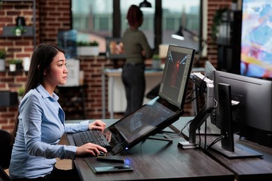 confident asian 3d model creator sitting at desk with multiple monitors while working on cgi. digital artist creating computer generated imagery for game development while in creative agency workspace