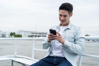 asian handsome male sit on ferry travel in the city for holiday trip. attactive young man backpacker tourist use mobile phone chatting message while sit on boat enjoy and happy spend time on vacation.