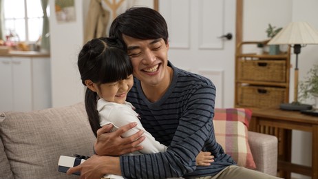 asian girl walking towards dad and showing him surprise gift on father’s day at home. the touched man giving his daughter a big hug on the sofa