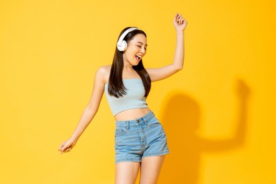 young pretty asian woman wearing headphones listening to music and dancing in the studio yellow color background